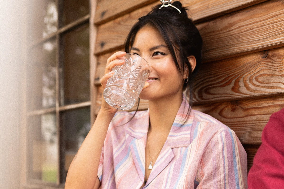 A woman sitting against a rustic wooden wall drinking from a clear glass mug.