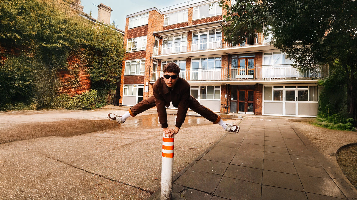 A young runner in sunglasses stretching against a street bollard on a wet pavement, a red-brick building behind him.