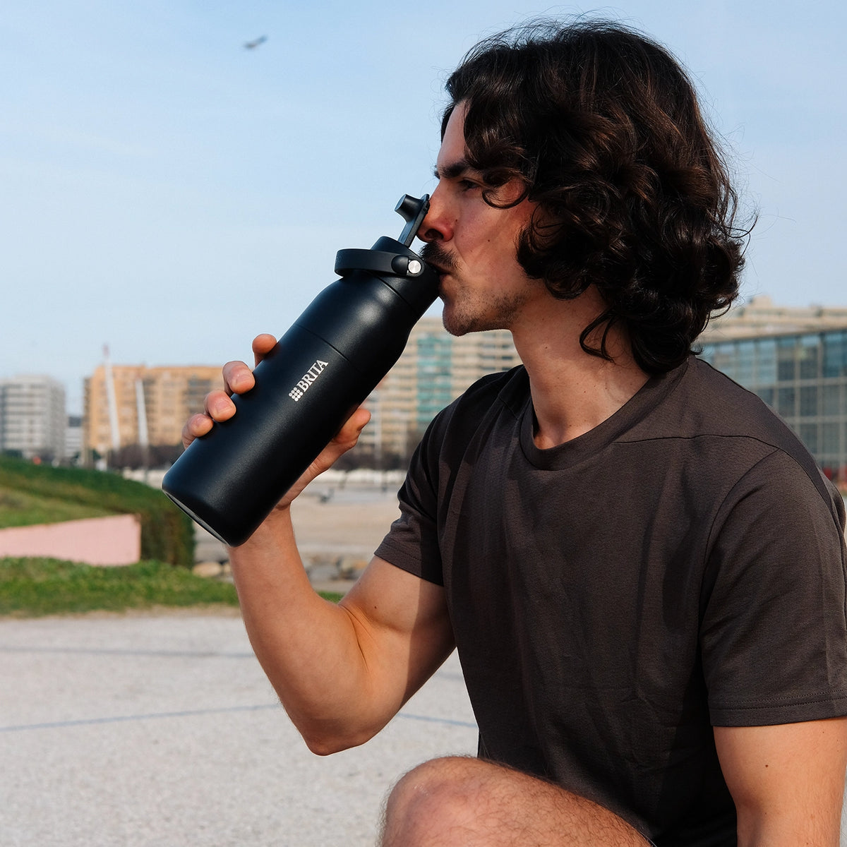 Person drinking from a black insulated bottle outdoors in an urban setting.
